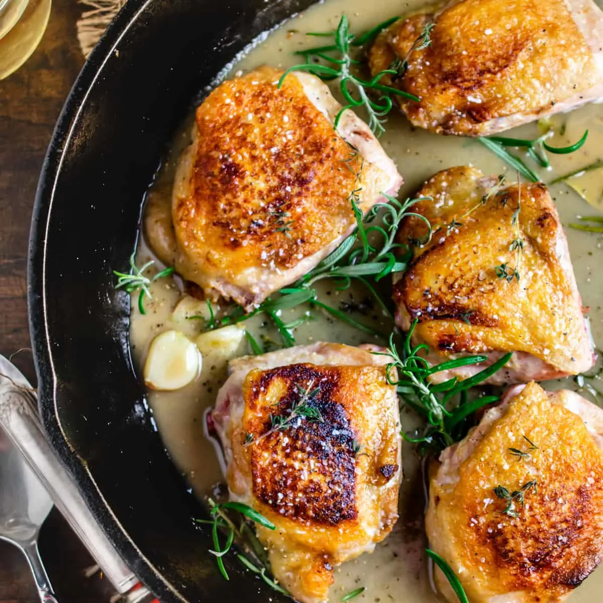 Seared chicken thighs in a cast iron pan with fresh herb sprigs and sauce.