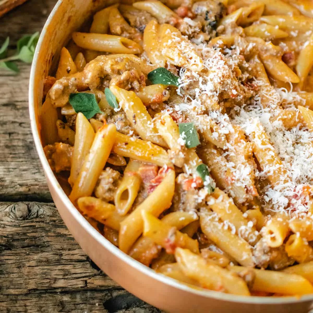 A skillet of penne pasta with vodka sauce, sausage and parmesan cheese on a rustic wooden back drop.