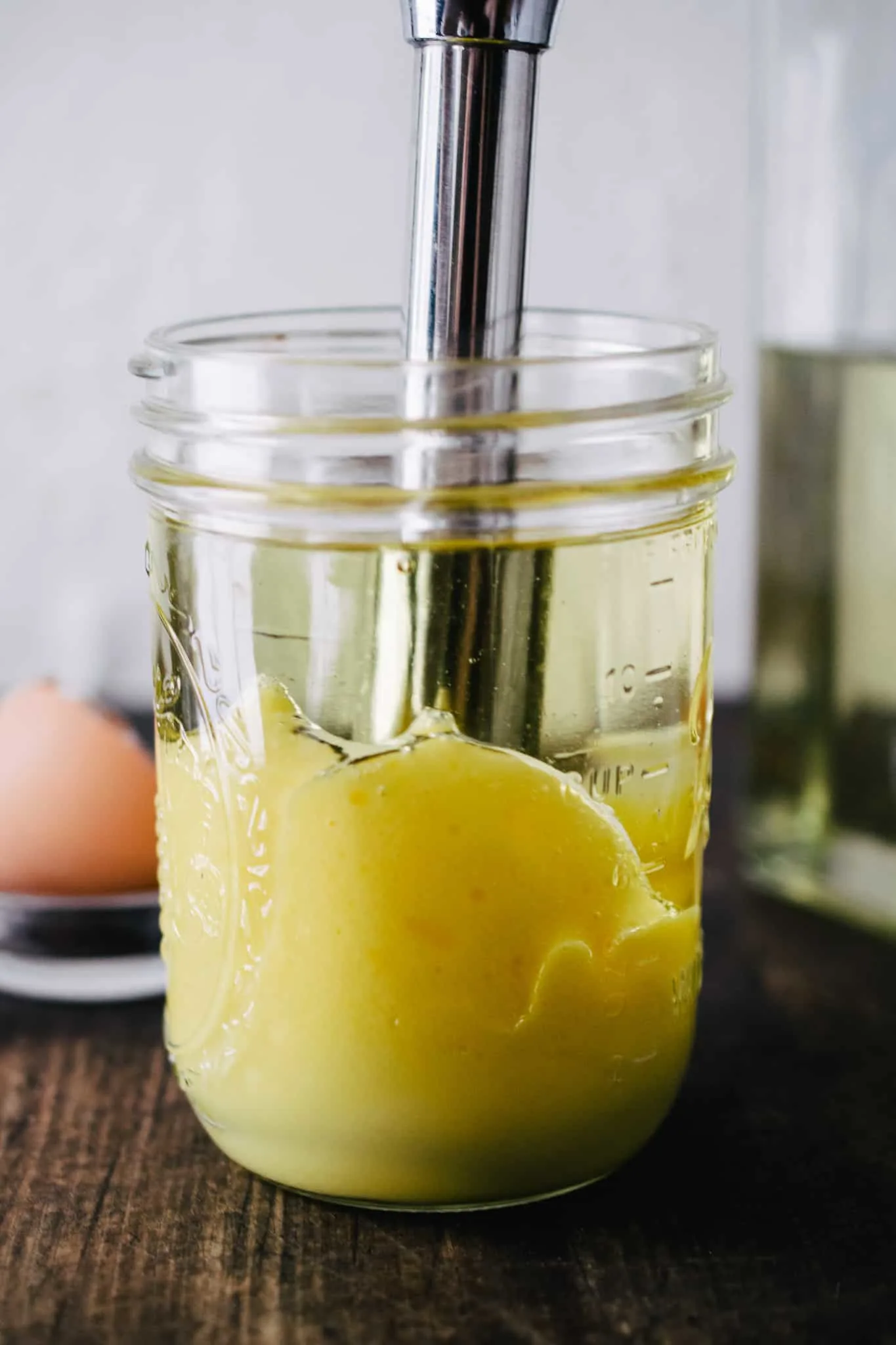 immersion blender in mason jar making homemade mayonnaise on dark wooden surface with white stone backdrop