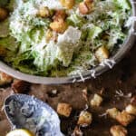 stainless steel bowl of tossed Ceasar salad, baking tray with homemade croutons, spoon and lemon wedge