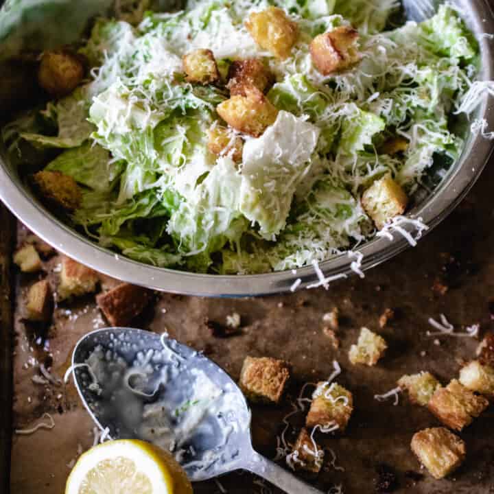 stainless steel bowl of tossed Caesar salad, with croutons, lemon wedge and spoon