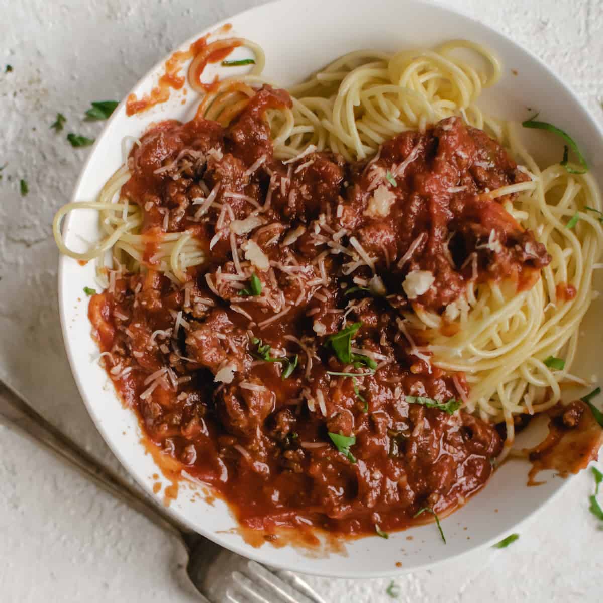 white bowl with spaghetti pasta, meat tomato sauce and torn fresh basil leaves next to a silver fork