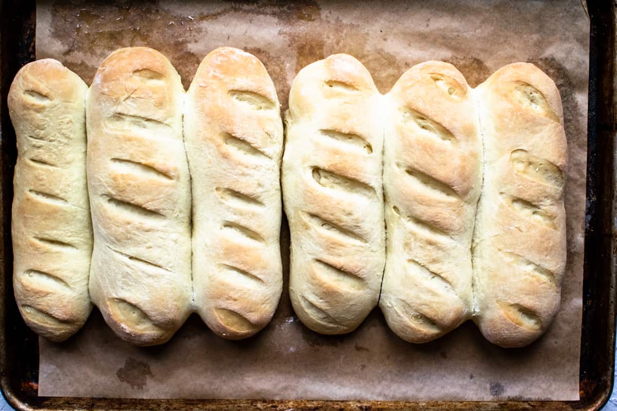 Six baked hoagie rolls on a baking sheet.