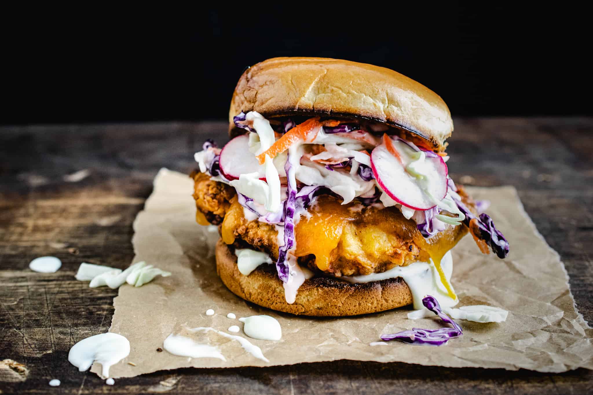 buttermilk fried chicken sandwich topped with cheddar and slaw, on brioche bun on wood surface with black background and messy drips scattered around