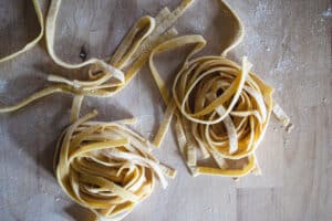 two homemade fettuccine pasta ribbons in nests on floured wood butcher block surface