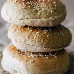 3 homemade sesame seed hamburger buns stacked on top of each other on brown paper surface and background