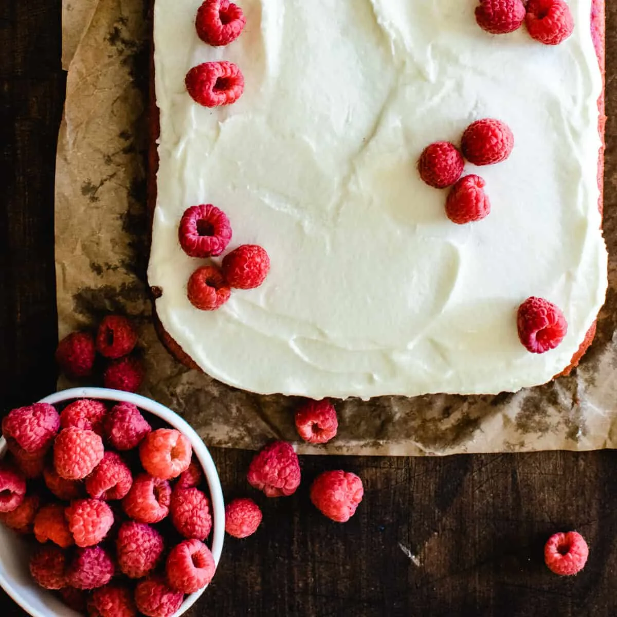 raspberry sheet cake on brown parchment paper with white frosting and bowl of fresh raspberries to decorate the top
