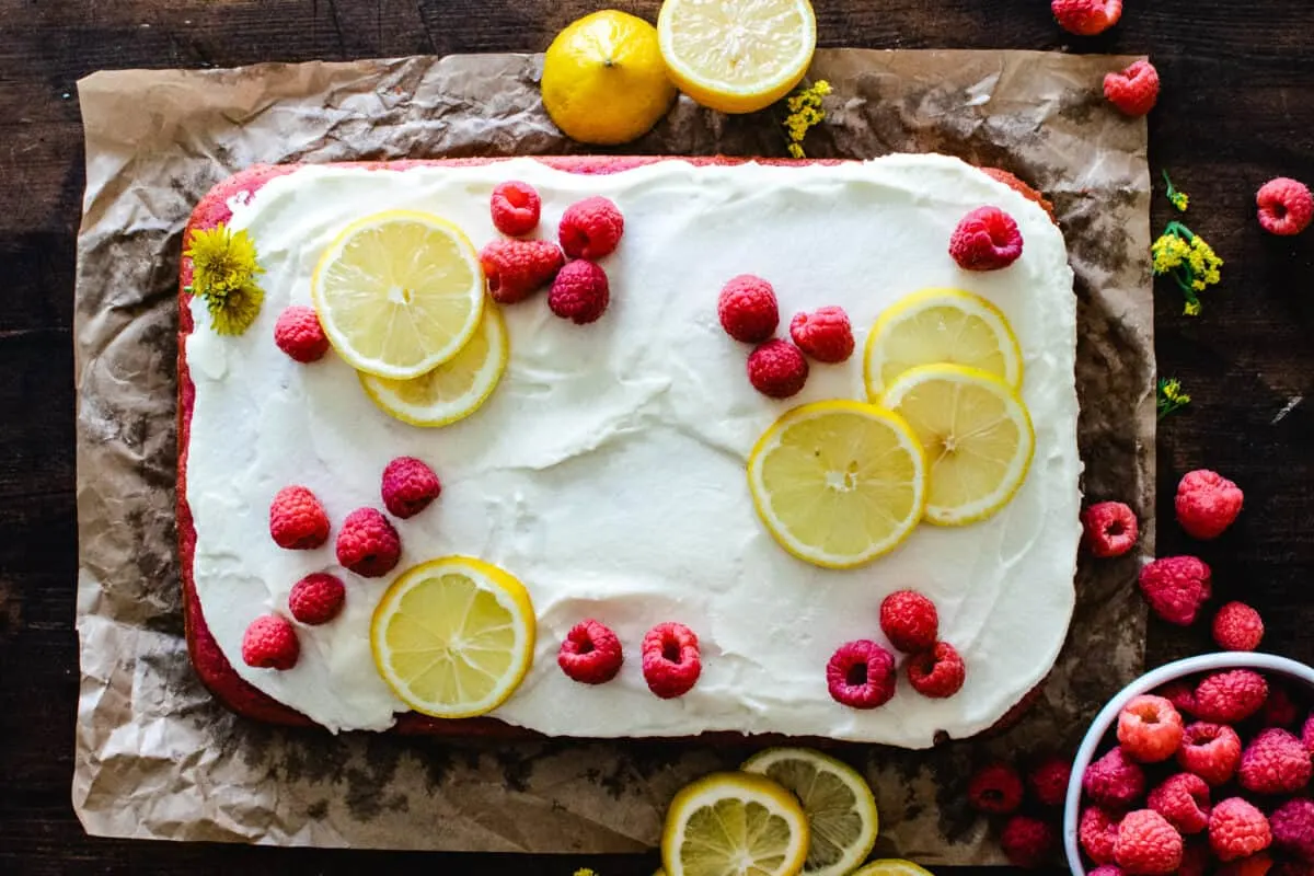 raspberry sheet cake with white frosting and fresh raspberries and lemon slices on brown parchment paper