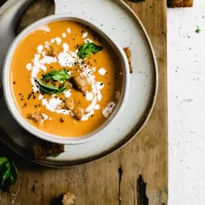 Bowl of tomato soup topped with fresh basil leaves and swirl of cream on plate and wooden board.