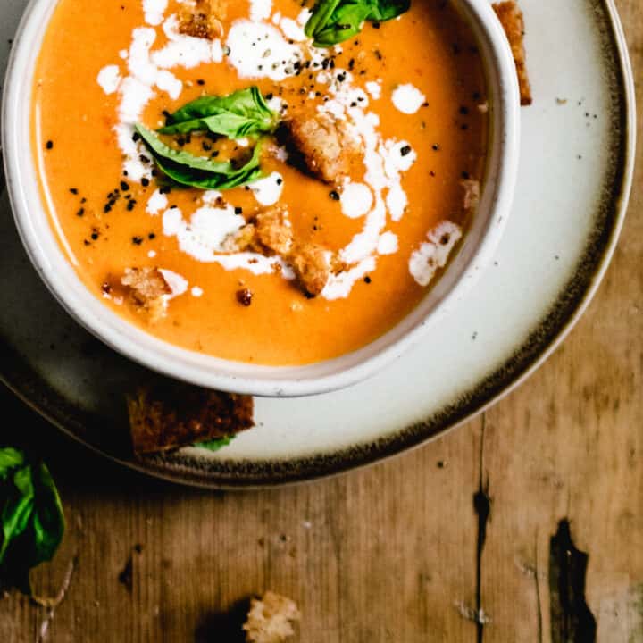 Bowl of tomato soup topped with fresh basil leaves and swirl of cream on plate and wooden board.