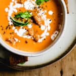 Bowl of tomato soup topped with fresh basil leaves and swirl of cream on plate and wooden board.