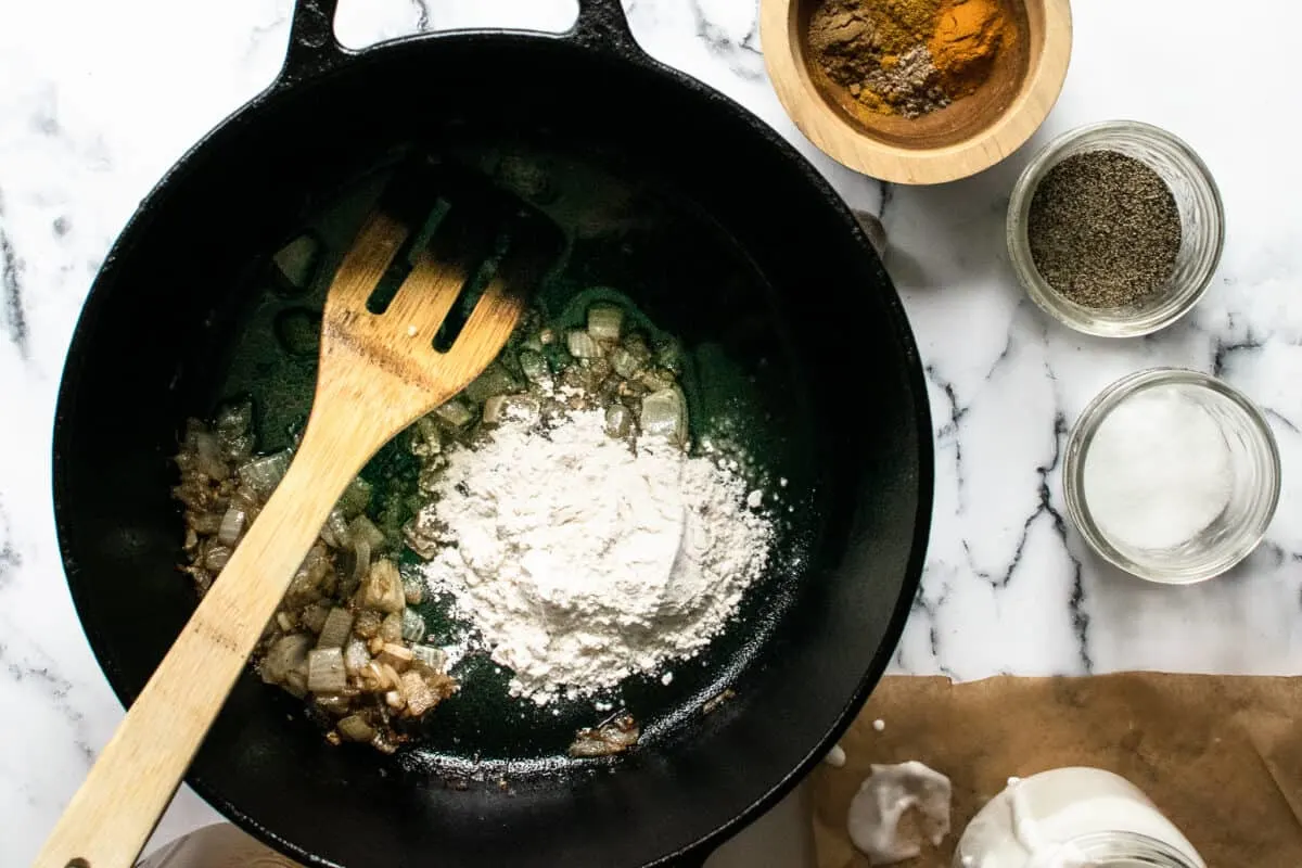 cast iron dutch oven sauteing onion and garlic with curry spices and flour, using a wooden spatula on a marble surface next to cup of coconut milk for adding later