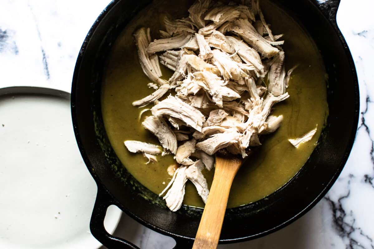 cast iron dutch oven with curry sauce and shredded chicken added about to be stirred up with a wooden spoon on a marble counter top