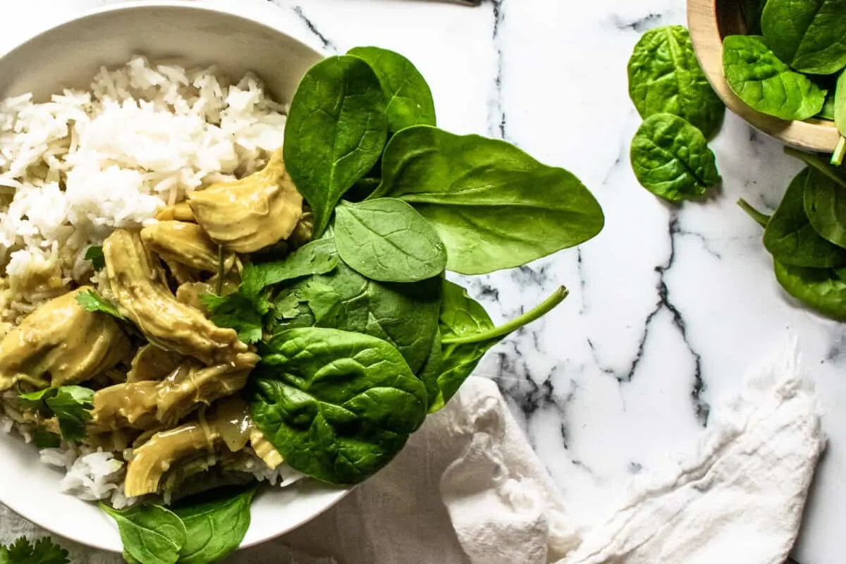 bowl of chicken curry and basmati rice served with a bed of fresh spinach leaves in a white bowl next to white napkin on marble counter and wooden bowl of extra fresh spinach