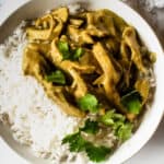 bowl of chicken curry with basmati rice in white bowl topped with cilantro on marble surface next to white fringe napkin, spoon and fork