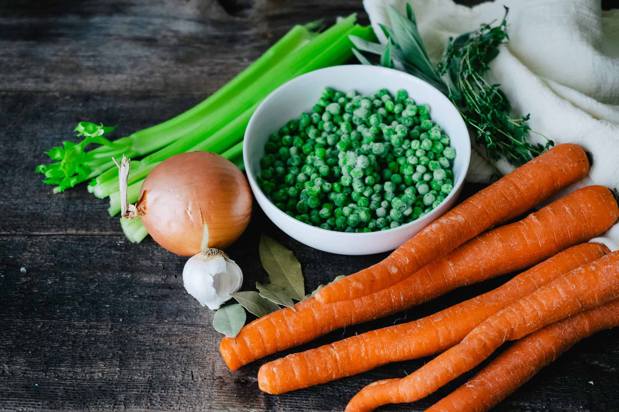 carrots, an onion, garlic cloves, celery and fresh herbs with a white bowl of frozen peas and white kitchen towel on a wooden surface