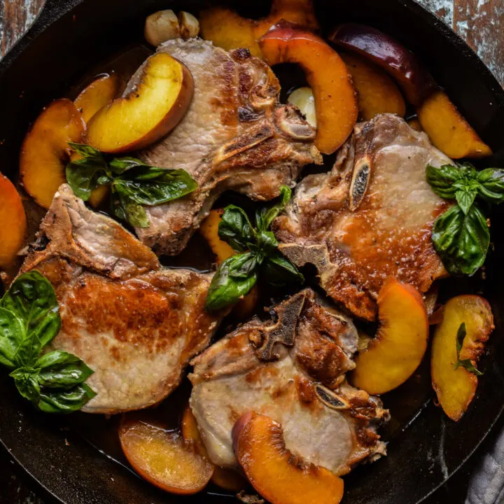 Bone in pork chops in a cast iron skillet with peaches and basil leaves.