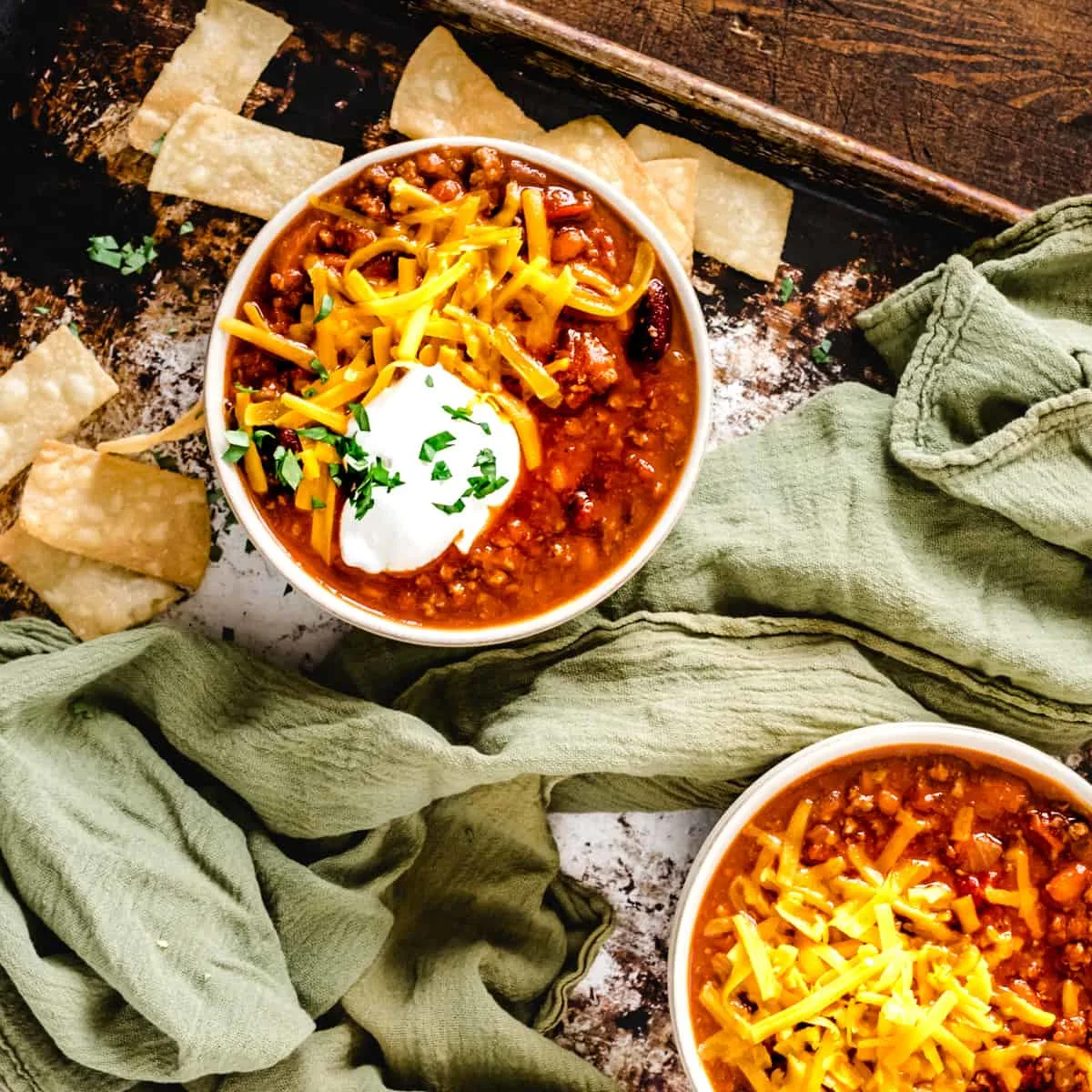 Two bowls of chili topped with sour cream, cheese and tortilla chips on a green kitchen towel.