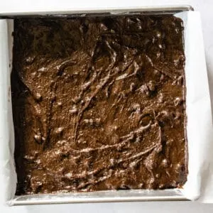 brownie batter being spread into a square, parchment paper lined baking pan