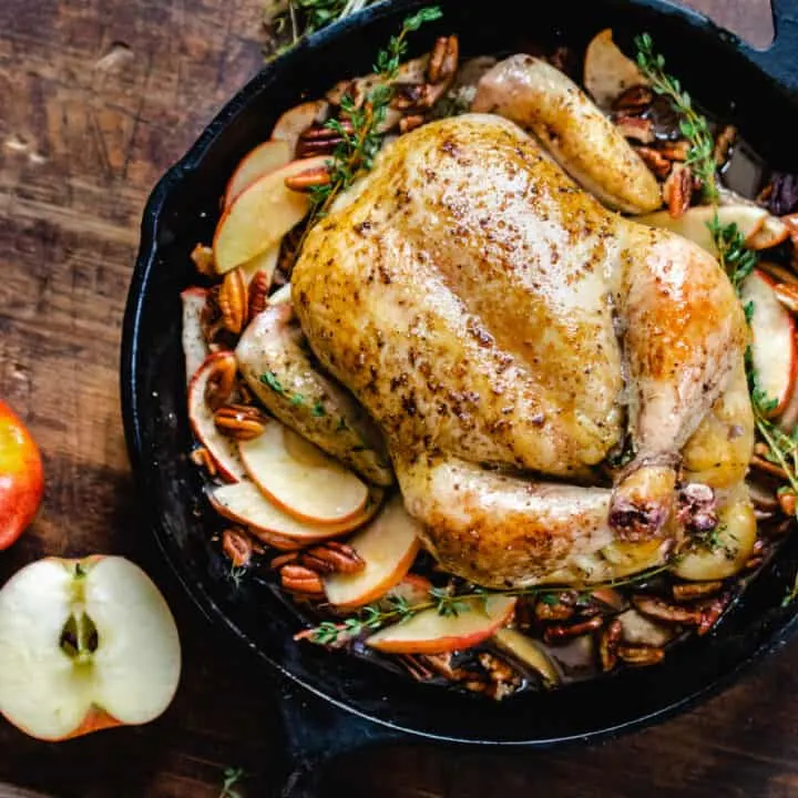 Whole, roasted chicken on a bed of sliced apples with sprigs of thyme in a cast iron skillet.