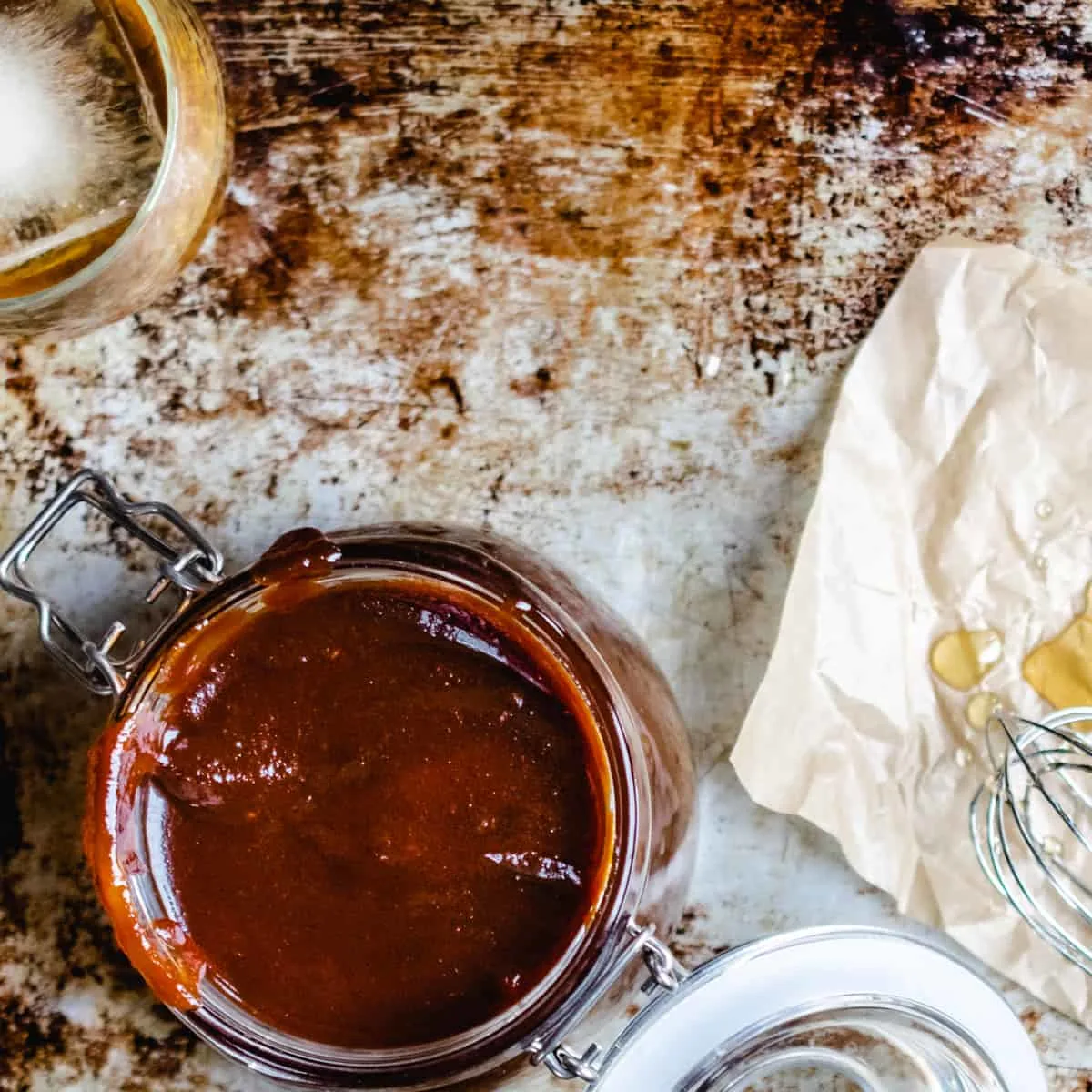 A jar of barbecue sauce next to a whisk with honey on it and glass of bourbon with ice.