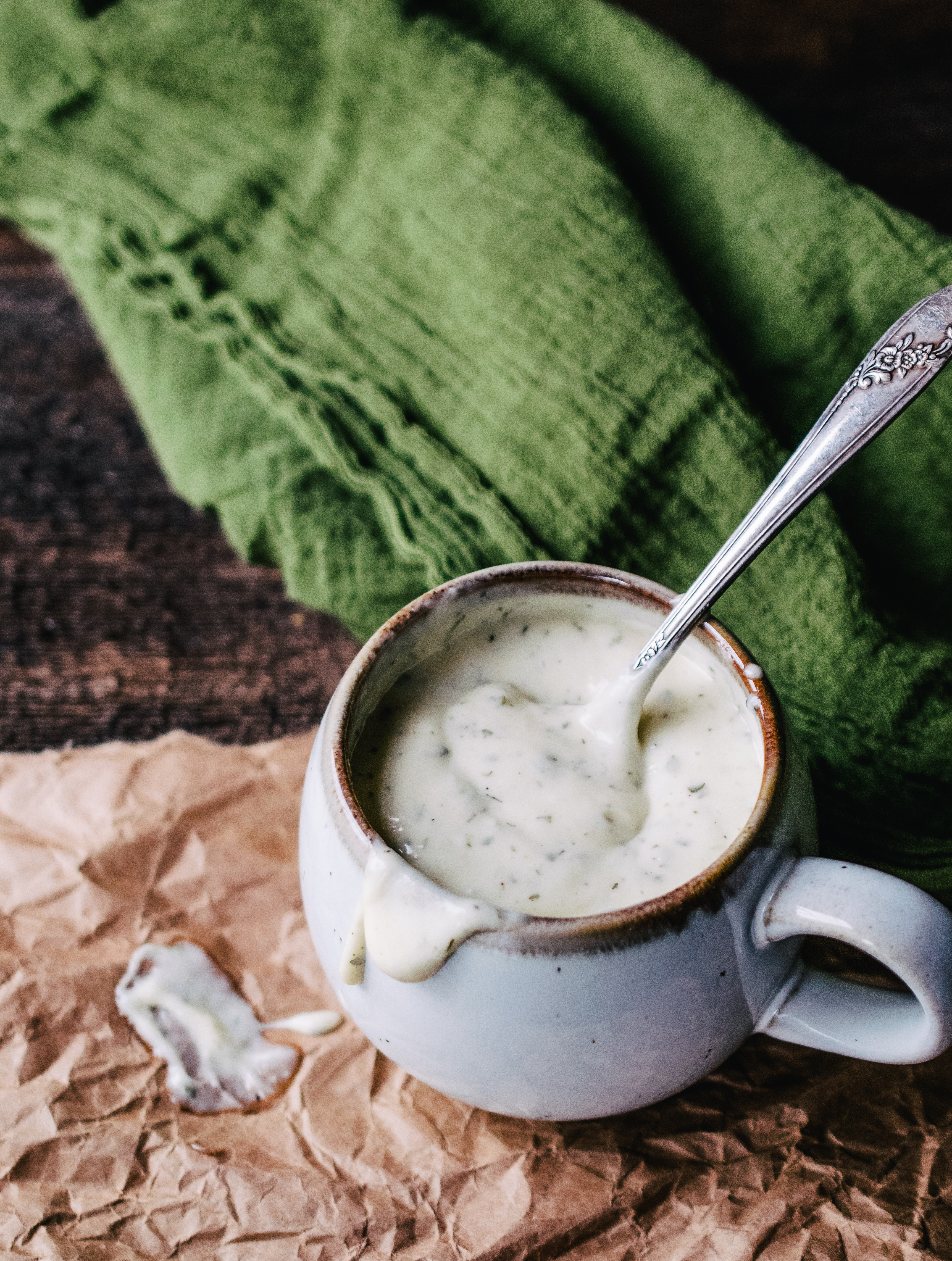 white speckled ceramic mug with brown rim containing homemade ranch dressing dripping down the side with antique silver spoon and green flour sack towel