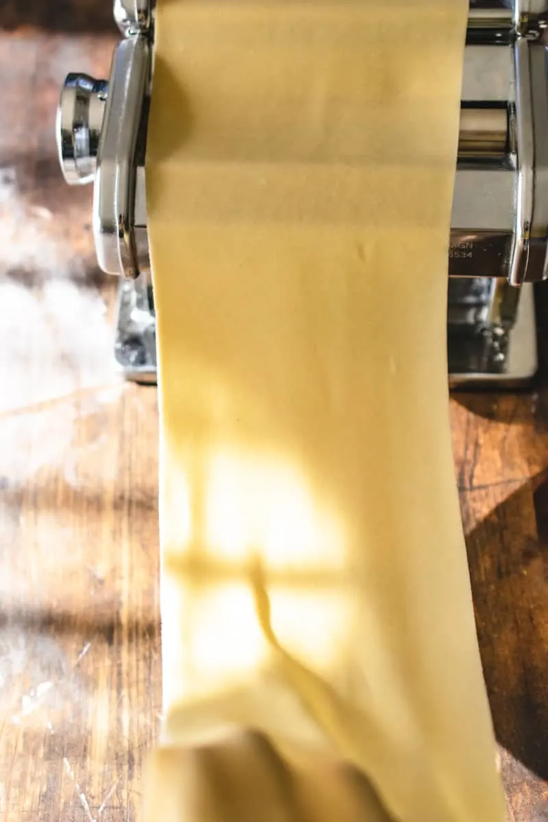 Long sheet of pasta dough being rolled through a hand held pasta roller.