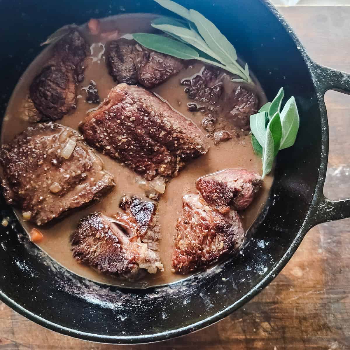Large chunks of beef in a braising liquid with herbs in a pot. 