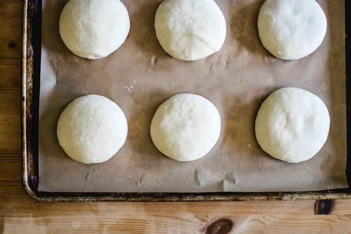 six raw hamburger buns on a baking sheet