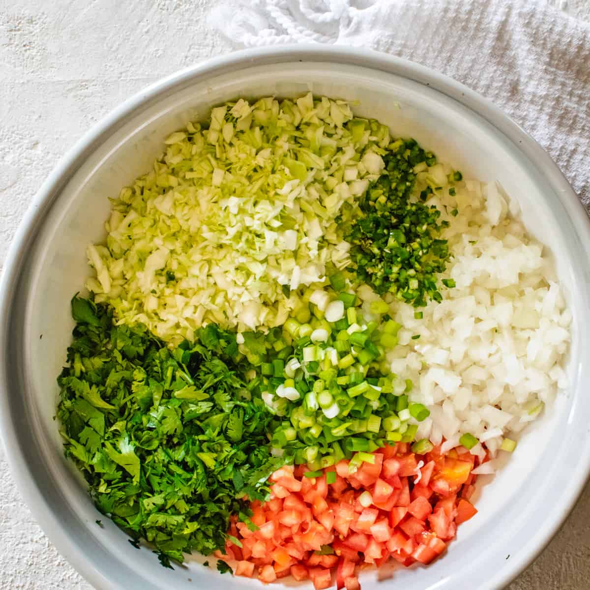 large bowl of finely chopped cabbage, tomato, onion, cilantro, Serrano, and scallions on white stone surface