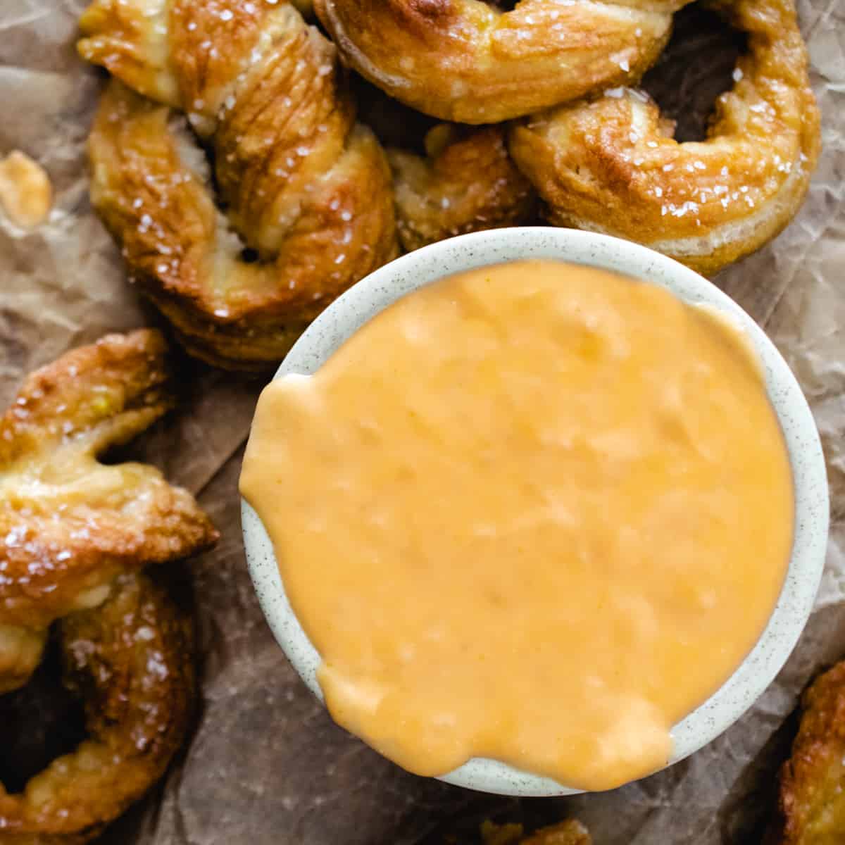 Bowl of creamy cheese dip surrounded by soft pretzels with salt on brown paper. 