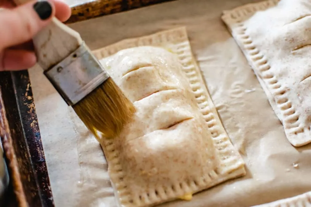 Brushing an uncooked ham and cheese pocket dough with beaten egg wash.