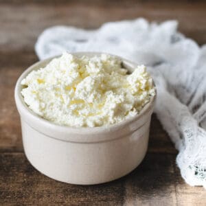 Beige stoneware ramekin full of ricotta cheese next to cheesecloth.