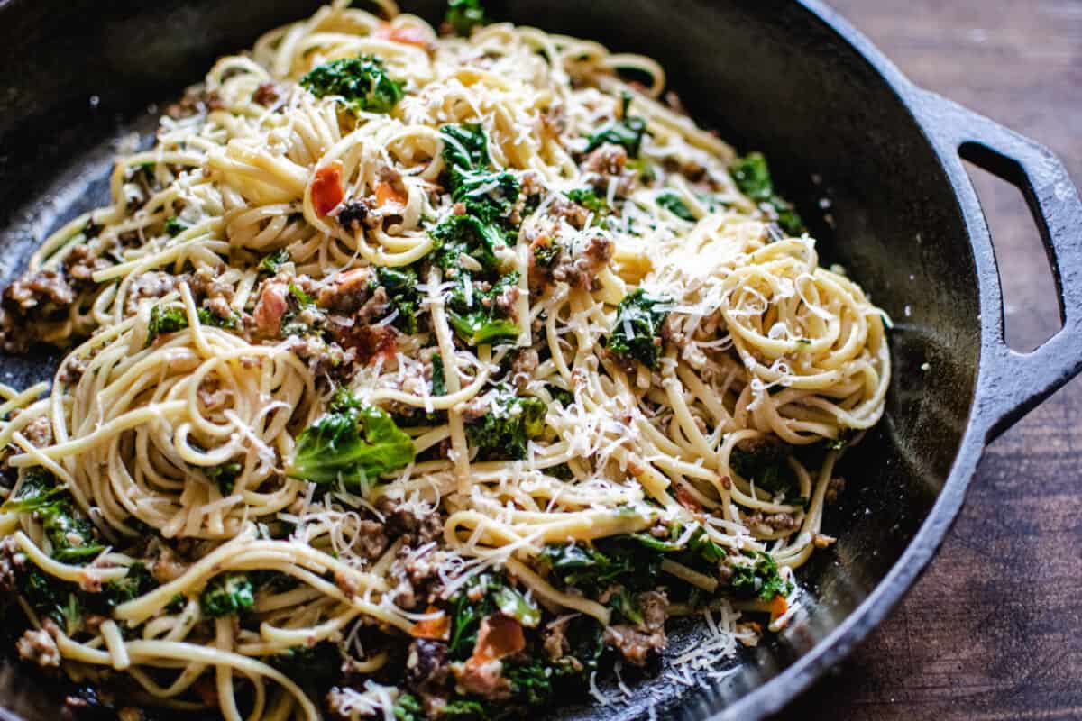 cast iron skillet of tossed ground sausage, kale and linguine pasta sprinkled with Parmesan cheese in a light wine cream sauce