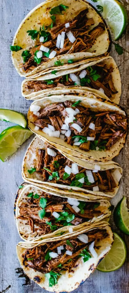 six corn tortillas with shredded beef, onion, cilantro and lime wedges on rustic wood board