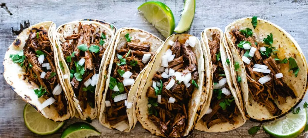 six tacos de barbacoa topped with cilantro and onion with lime wedges on rustic wood