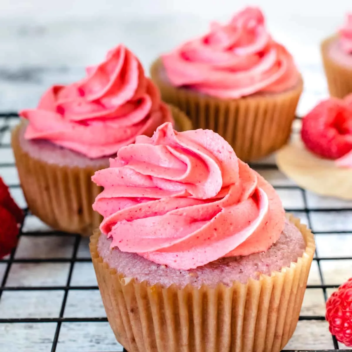 A pink cupcake with pink frosting surrounded by fresh raspberries.