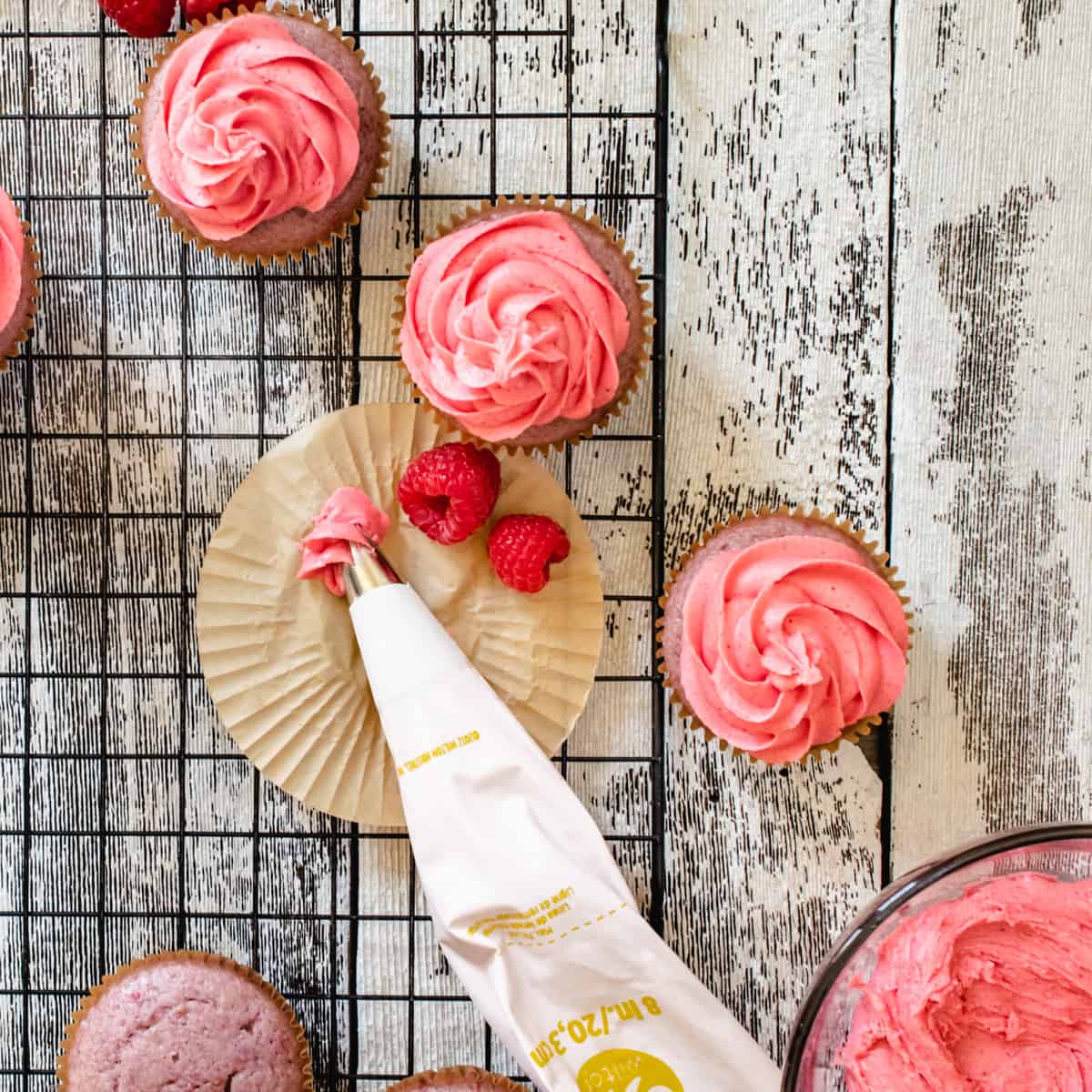 Piping bag with pink frosting, raspberries and cupcakes for decorating.