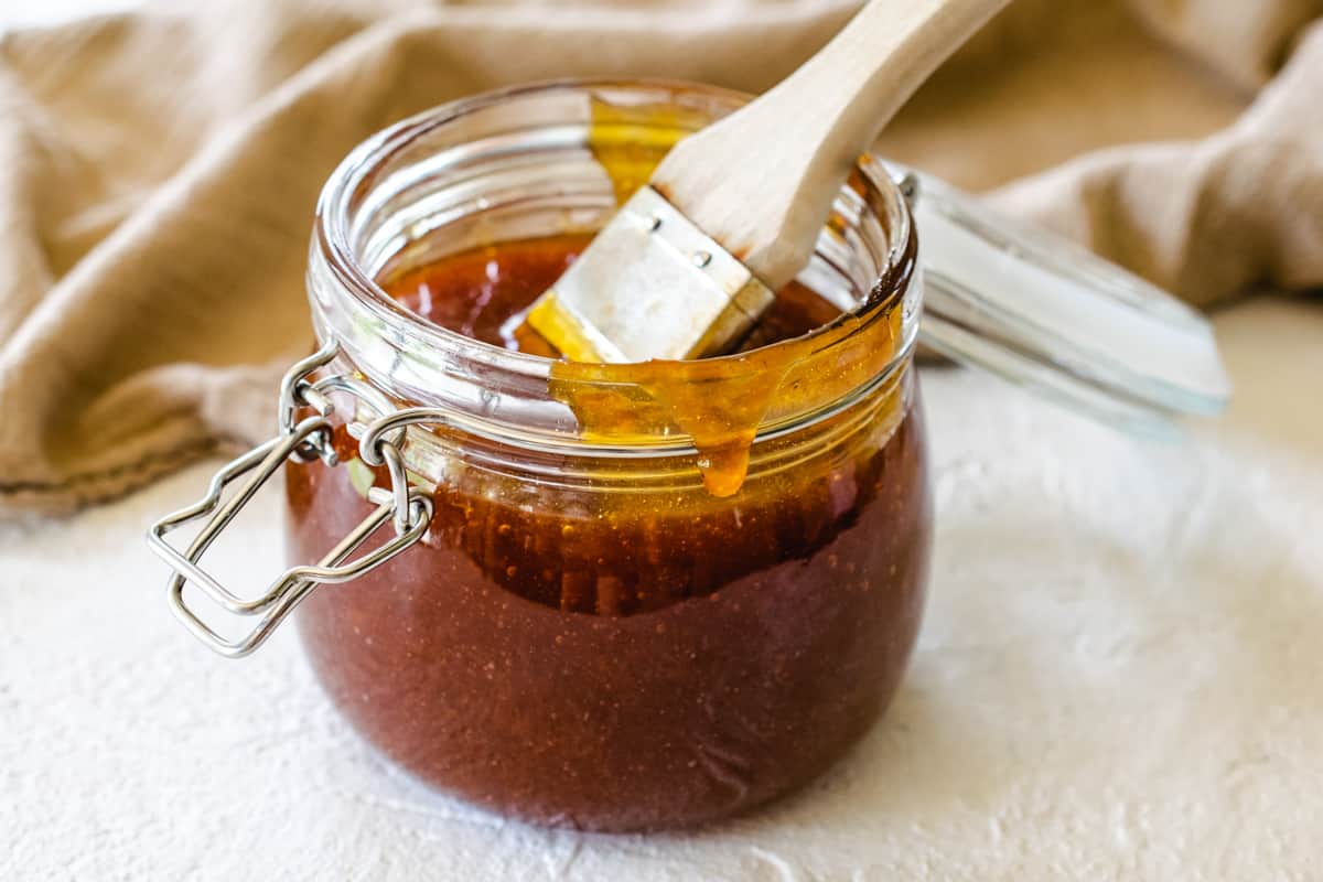 glass jar with hinged lid of apricot barbecue sauce and basting brush by taupe towel on white stone surface