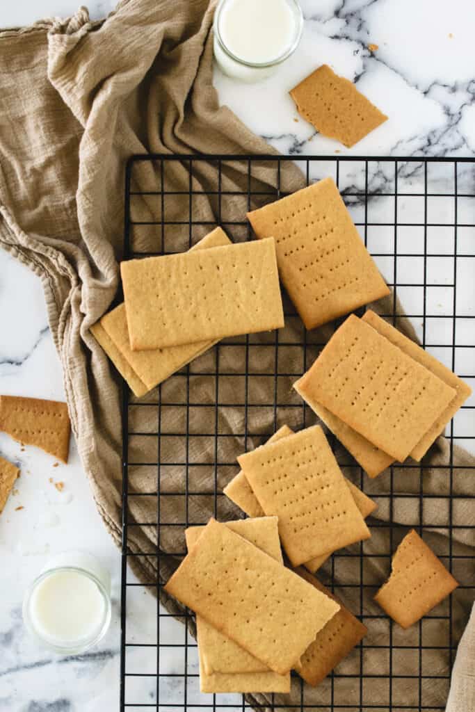 piles of graham crackers over a taupe colored towel on a cooling rack with two glasses of milk