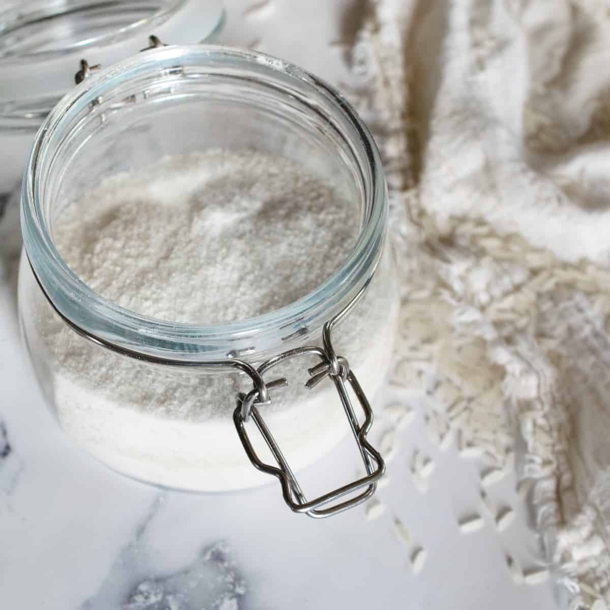 glass hinged jar with rice flour next to grains of rice sprinkled over a white fringe towel