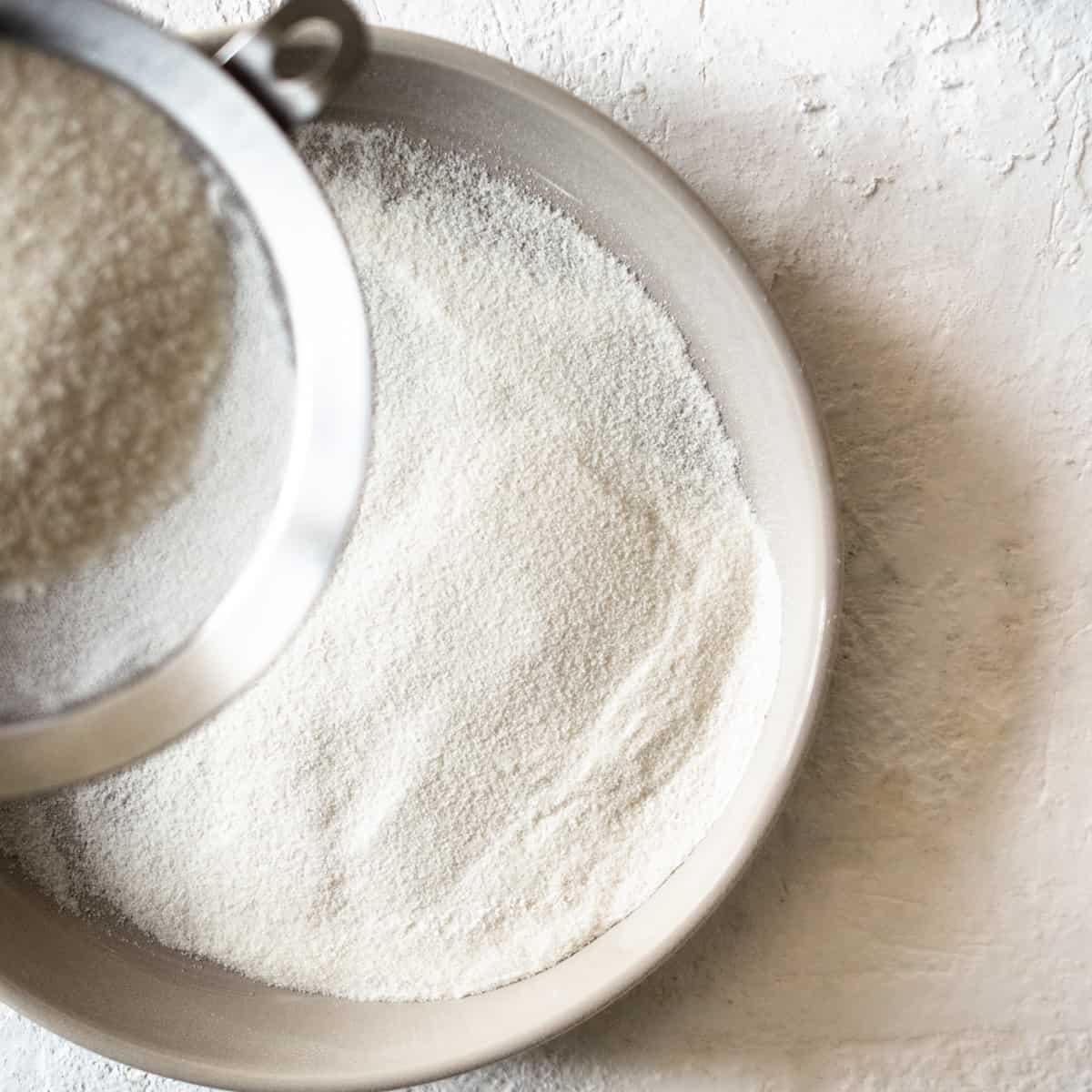 Rice flour being passed through a metal mesh strainer into a shallow bowl.