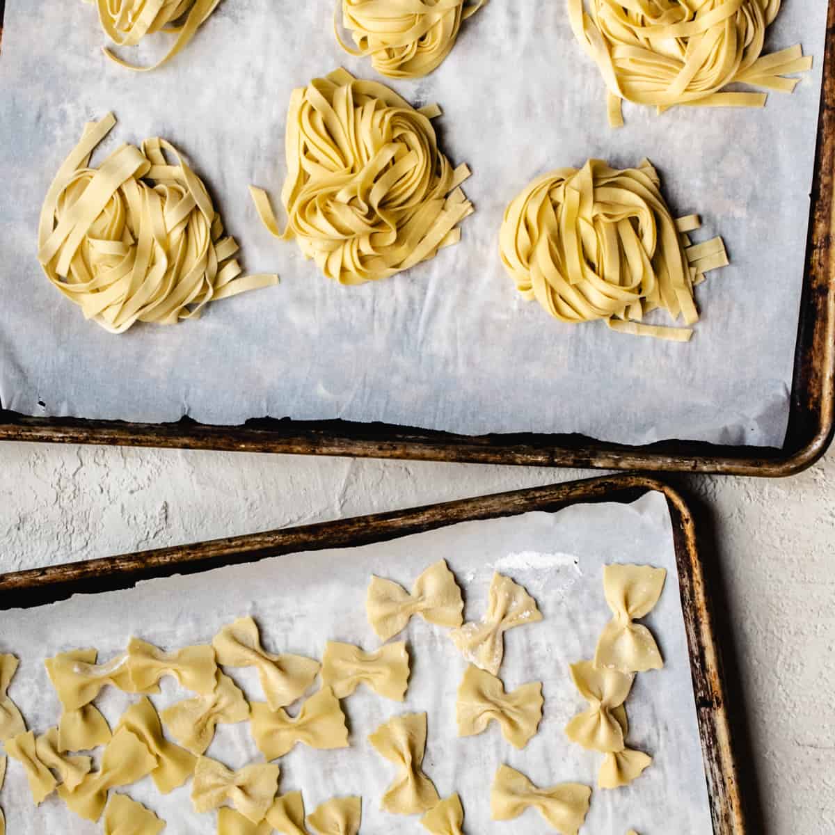 Parchment paper lined baking sheets with fettuccine and farfelle pasta shapes.