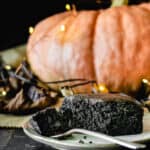 Piece of black chocolate cake with a bite taken out in front of decorative lights and a pumpkin.