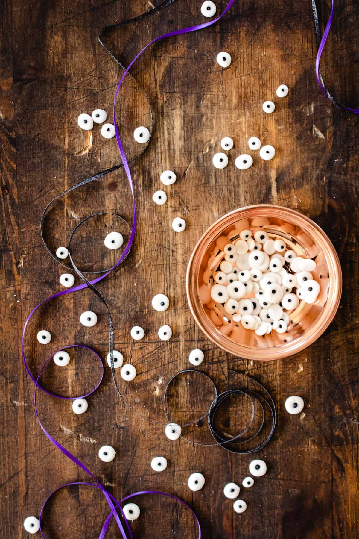 Candy eyeballs in copper dish and scattered on wood cutting board with black and purple ribbon. 