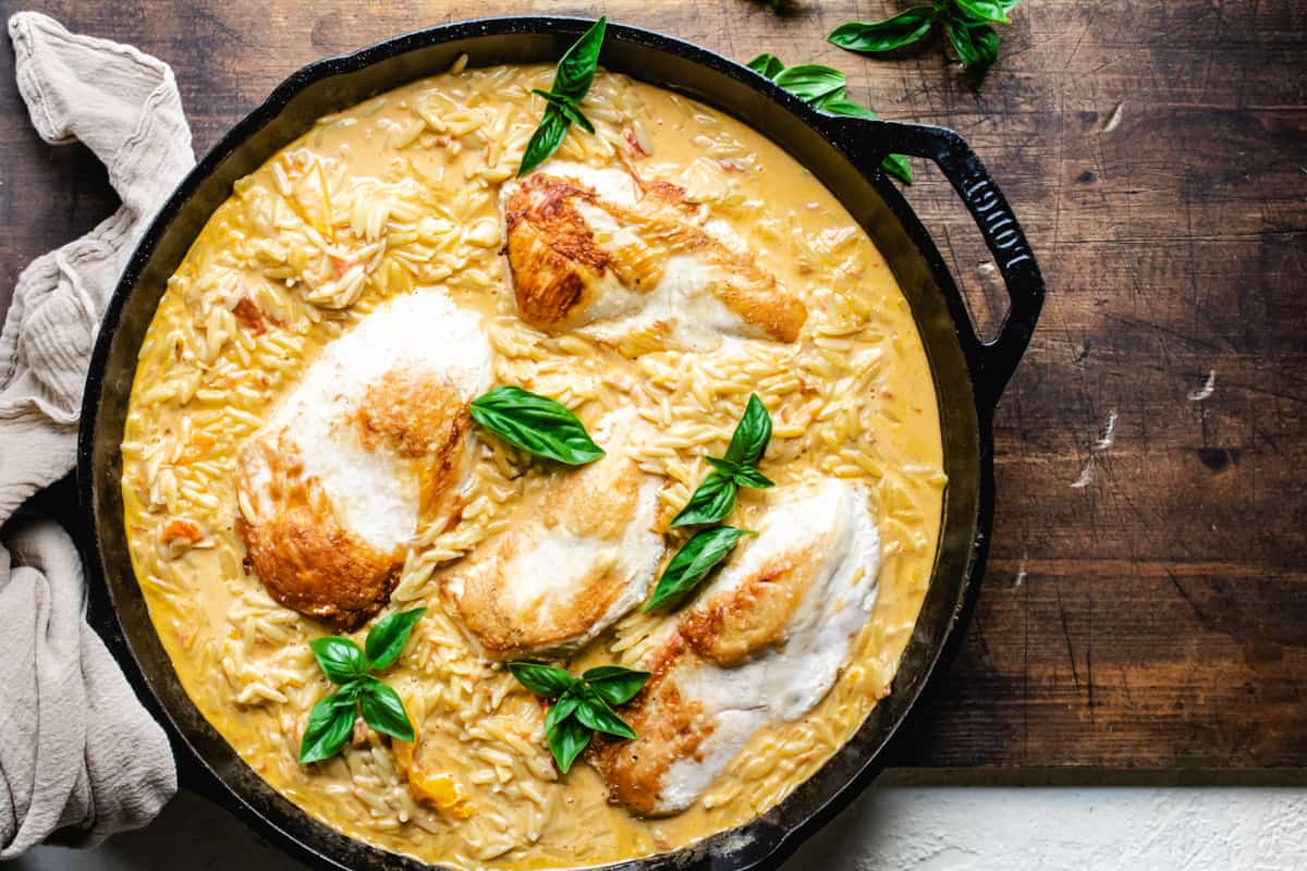 Cast iron skillet with creamy orzo, four chicken breasts, and fresh basil leaves on wooden cutting board. 