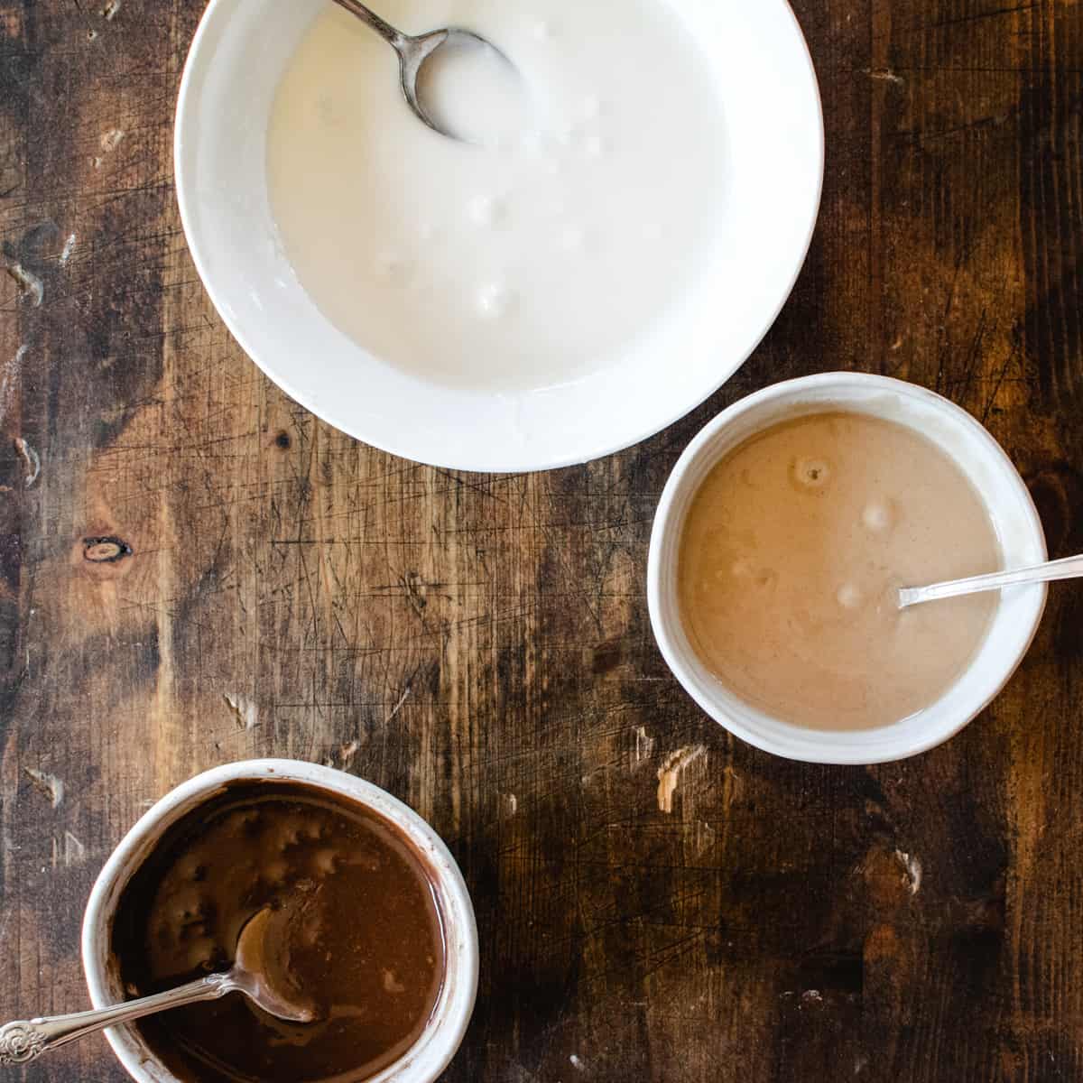 Three bowls of icing, one vanilla, one chocolate and one cinnamon on a wood surface. 