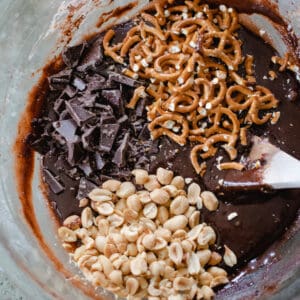 Bowl of brownie batter with piles of pretzels, peanuts and chocolate bar pieces.