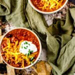 Two bowls of chili with sour cream, cheese and cilantro on a green kitchen towel.