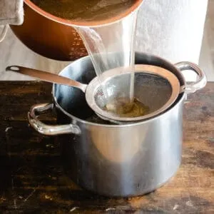 Pouring beef broth through a mesh screen into a large pot.